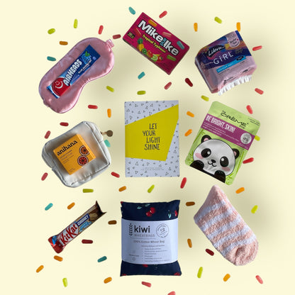 Contents of gift box displayed in the gift box. Kiwi wheat bag, gratitiude journal,glow lab face mask, Mike and Ike candy, kit kat, little purse, airhead candy, eye mask, libra teen pads, fluffy socks and anihana shower steamer.