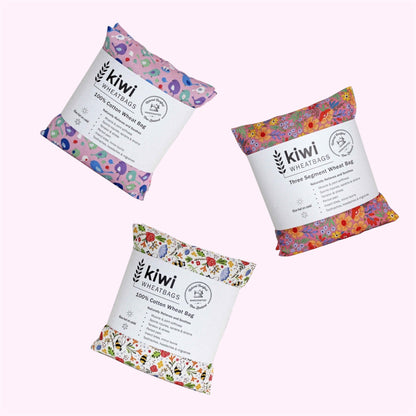 Three packaged wheat bags from kiwi WHEATBAGS arranged on a pale pink background. Each bag has a white paper label with black text and is wrapped in a colorful floral-patterned fabric.